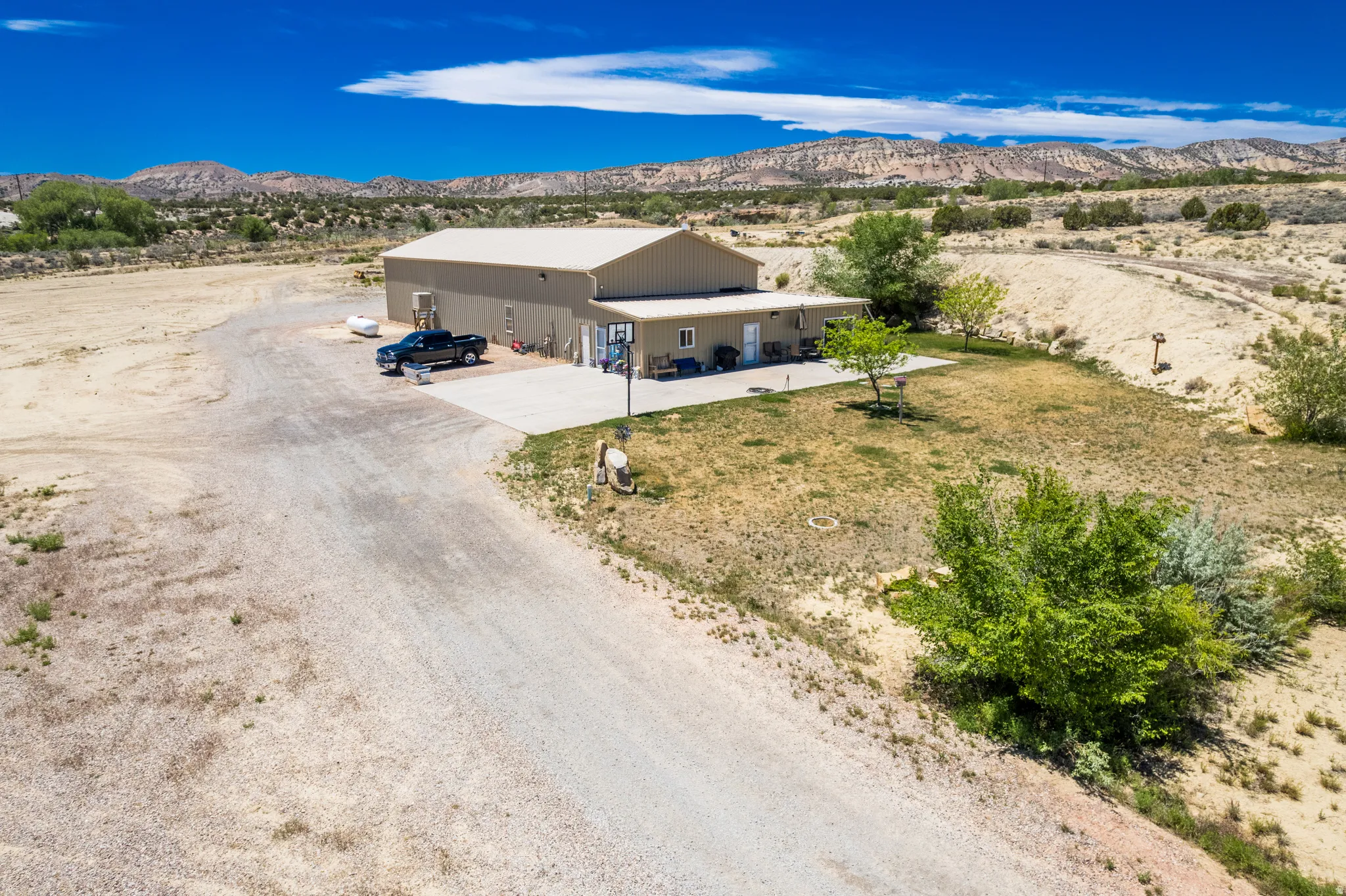 Expansive property featuring a large tan-colored building with a light-colored roof, a gravel driveway, a concrete patio, and scattered green trees