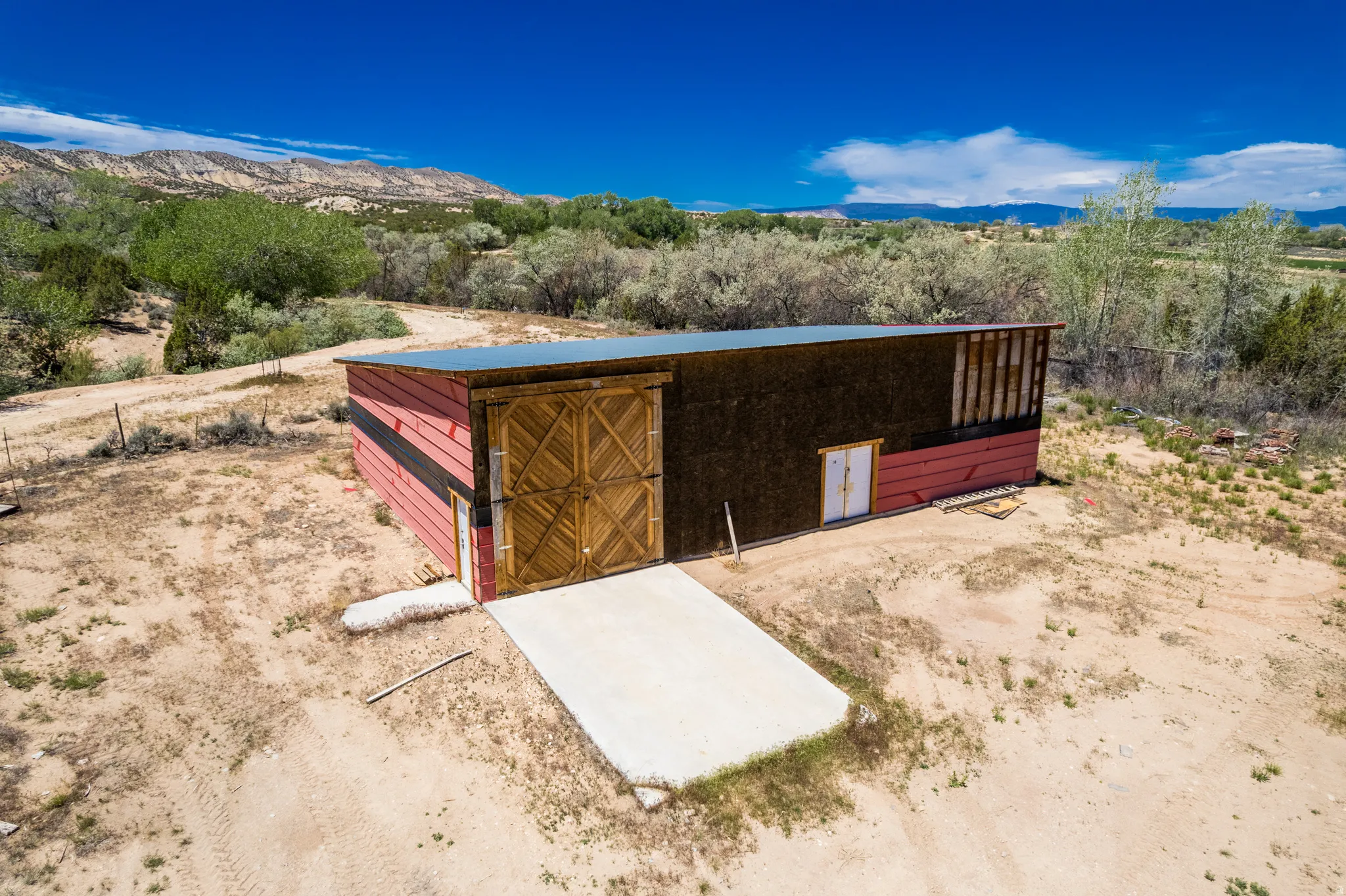 Barn structure featuring a red and dark wood exterior, large double wood doors with a diagonal pattern, and a concrete pad