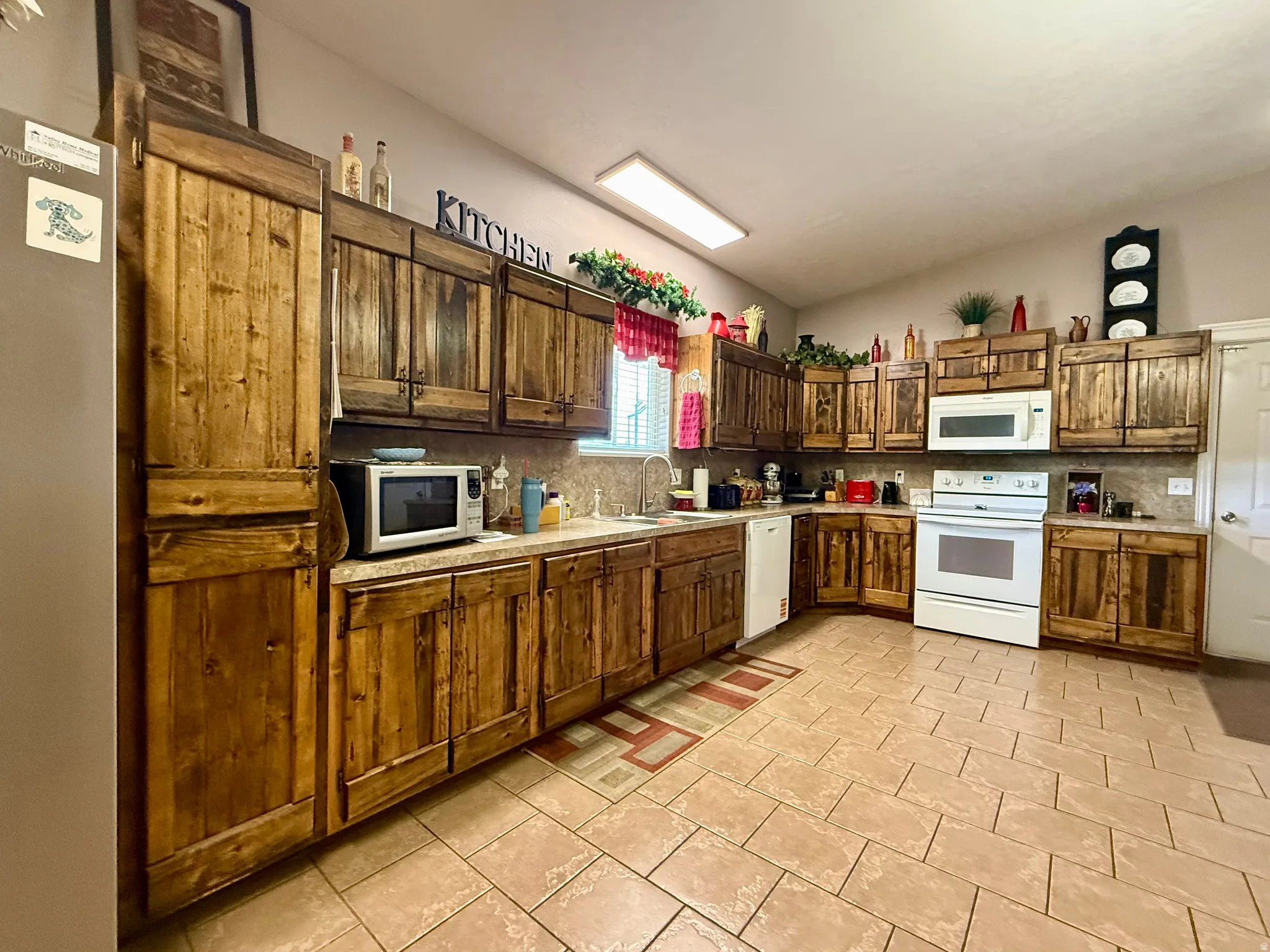Kitchen featuring wood-panel cabinetry, light-toned tiled flooring, white appliance suite, and a countertop with integrated sink