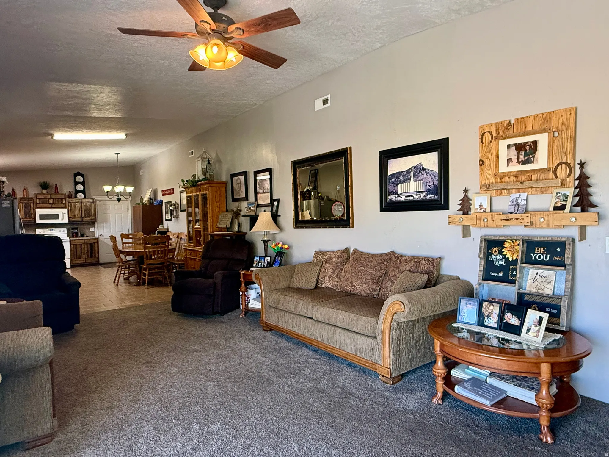 Open-concept living area featuring a ceiling fan with light fixture, carpet flooring, a dining area with a chandelier, and a kitchen with wood-finish cabinetry and built-in microwave