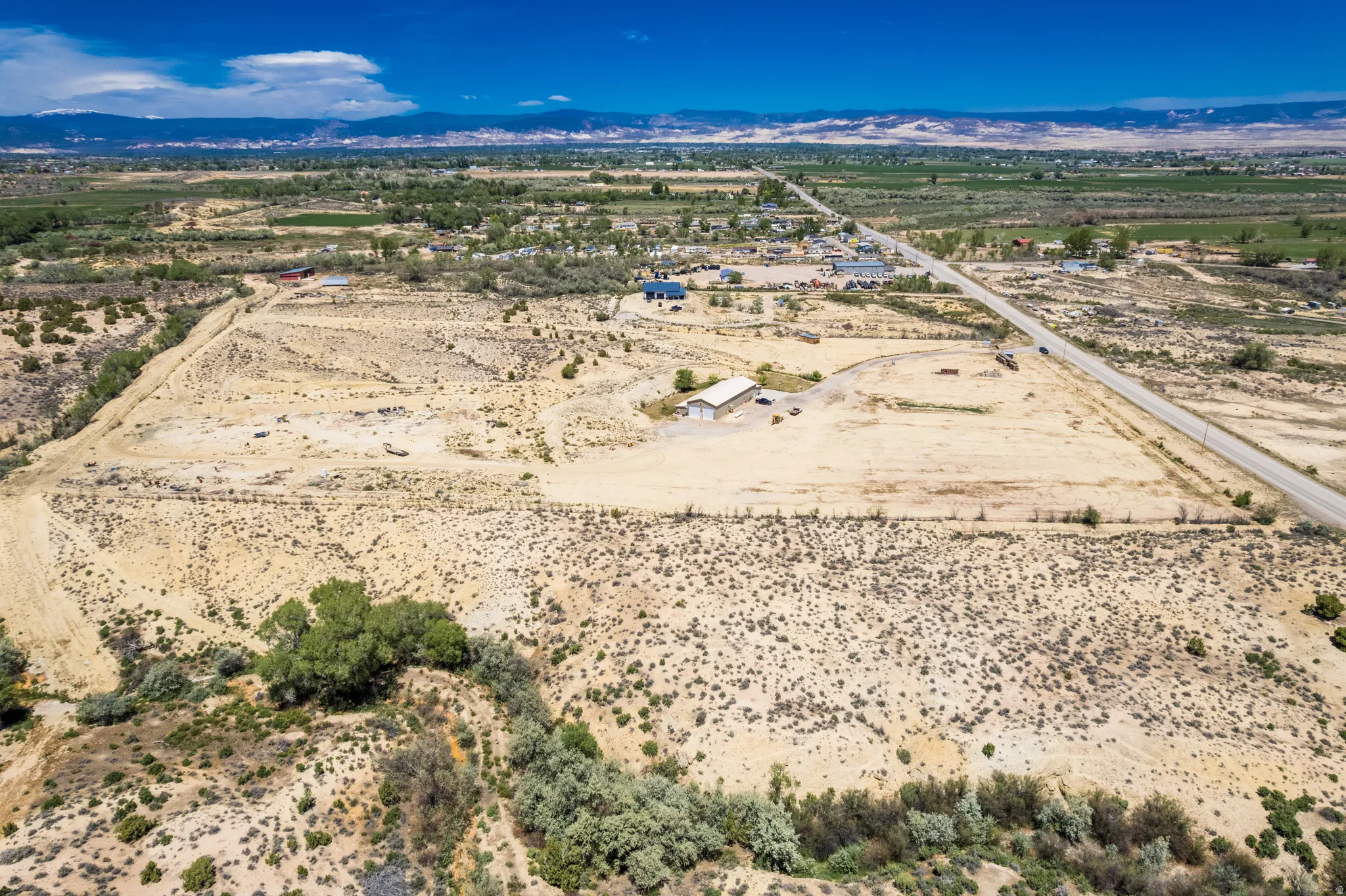 Expansive desert landscape featuring a paved road, scattered vegetation, a building structure with a light-colored roof, and distant mountains under a clear sky