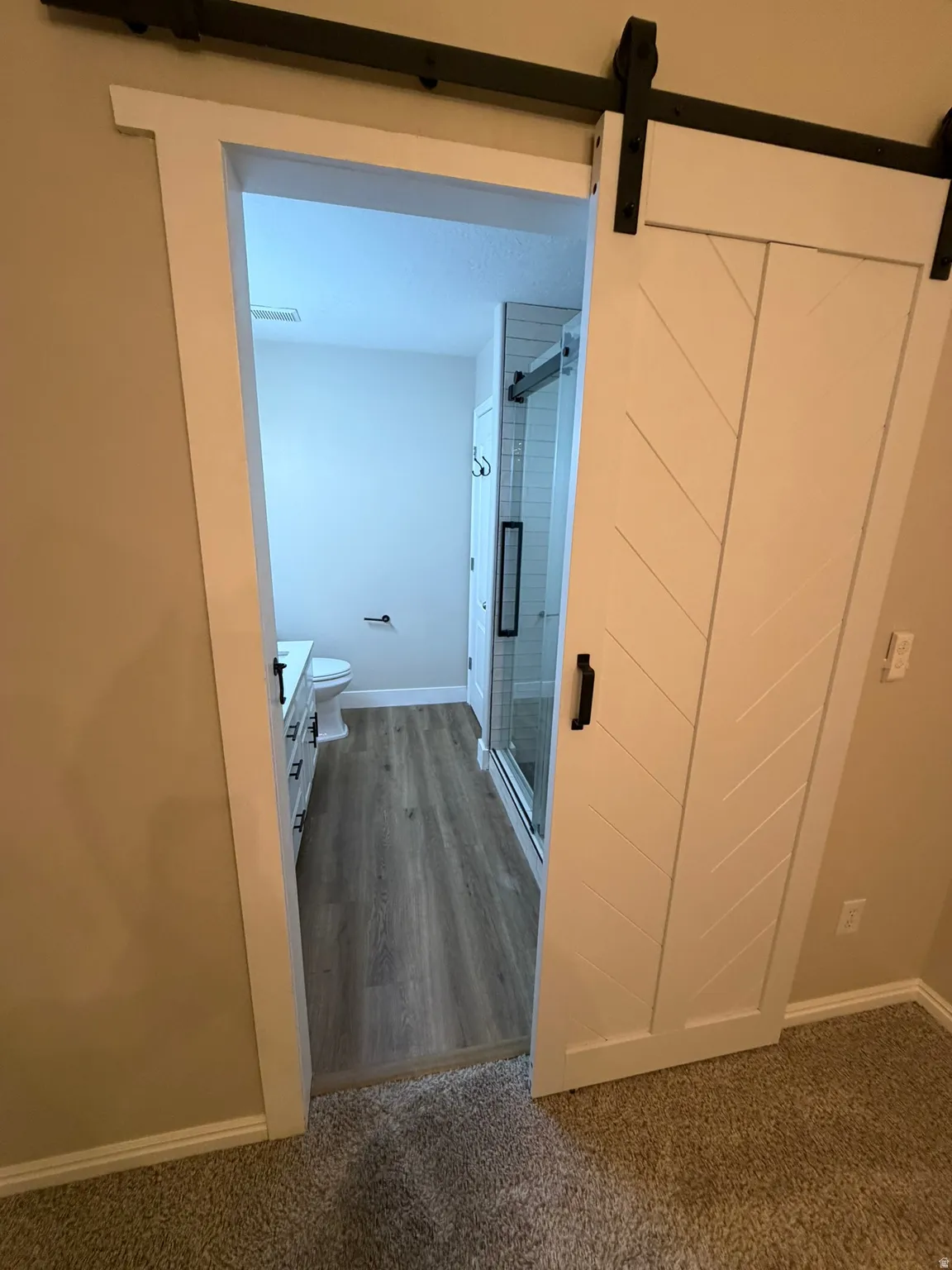 Modern farmhouse sliding barn door with black hardware, opening to a bathroom featuring wood-finish flooring, a white vanity with drawers, and a clear glass shower enclosure