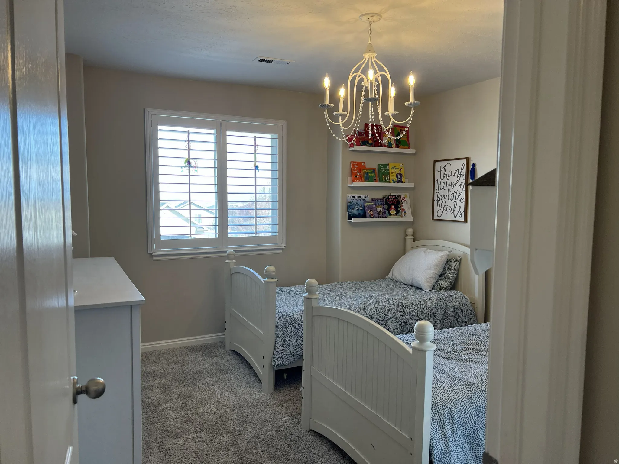 Neutral-toned room featuring a window with white plantation shutters, gray carpet flooring, and a decorative chandelier