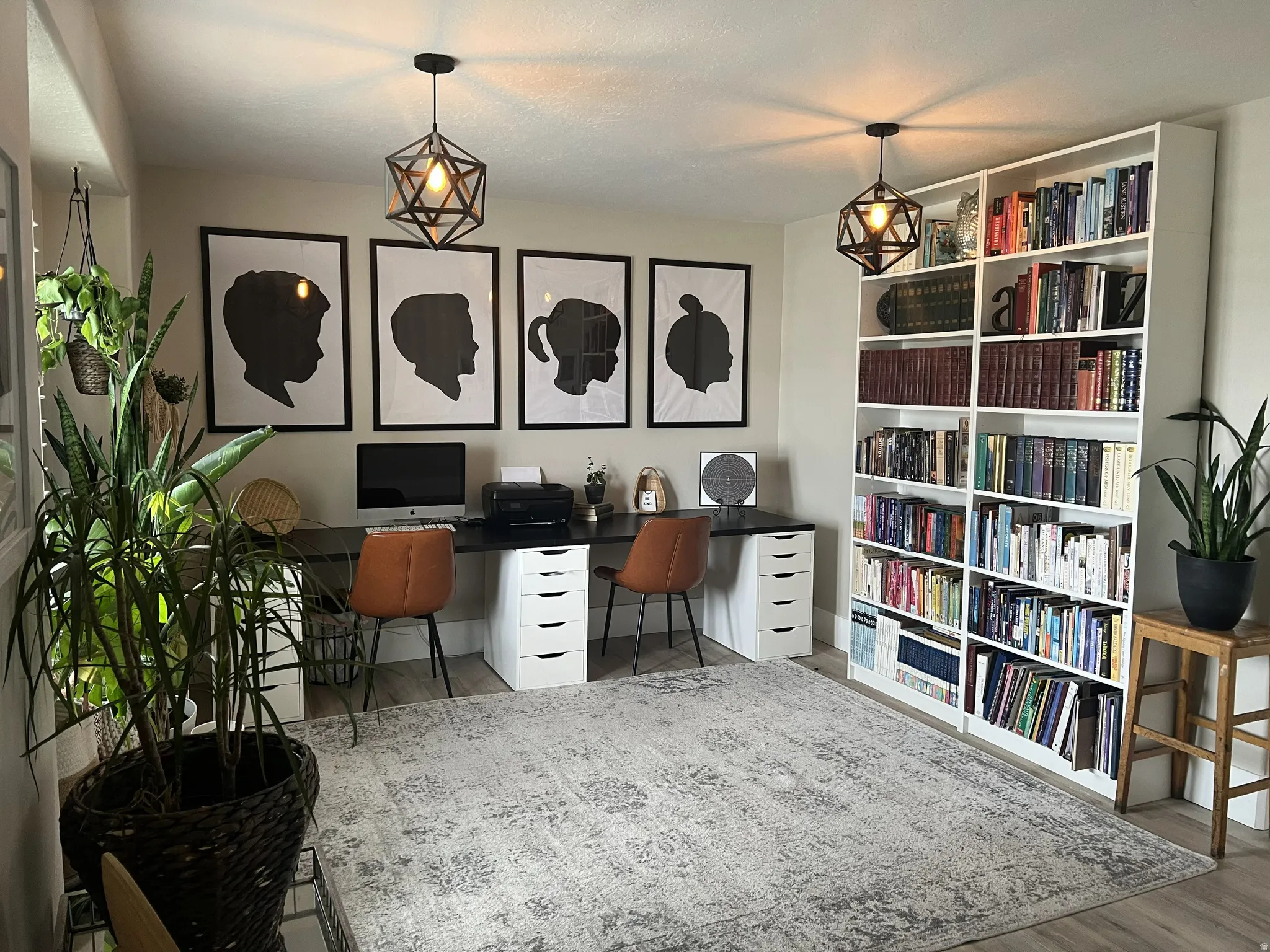 Dedicated office space featuring two geometric pendant lights, a full-wall built-in bookcase, wood-finish flooring, and ample desk space with integrated drawer units