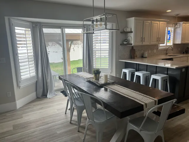 Open-concept dining area with wood-finish flooring, a contemporary linear chandelier, and bay windows featuring plantation shutters