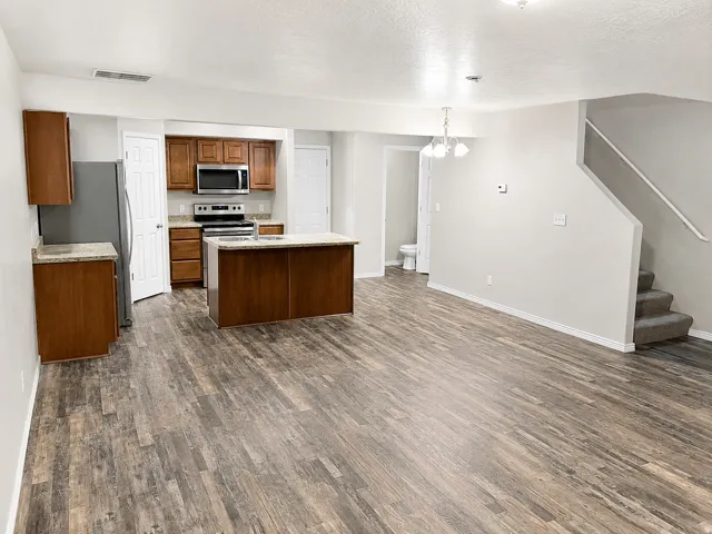 Open-concept living area featuring wood-finish flooring, a kitchen island with stone-look countertops, stainless steel appliances, and a carpeted staircase