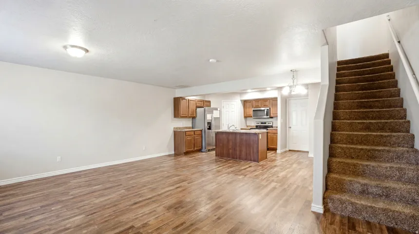 Expansive room featuring wood-finish flooring, a kitchen with wood cabinetry and stainless steel appliances, a central kitchen island, and a carpeted staircase
