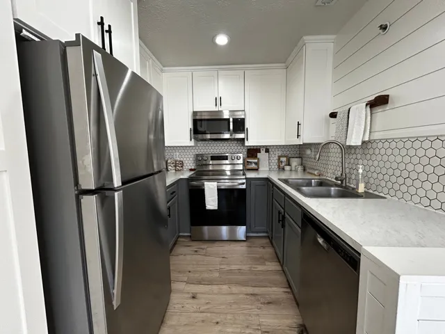 Galley kitchen featuring stainless steel appliances, two-tone cabinetry with black hardware, hexagon tile backsplash, light wood-finish flooring, and light-toned countertops