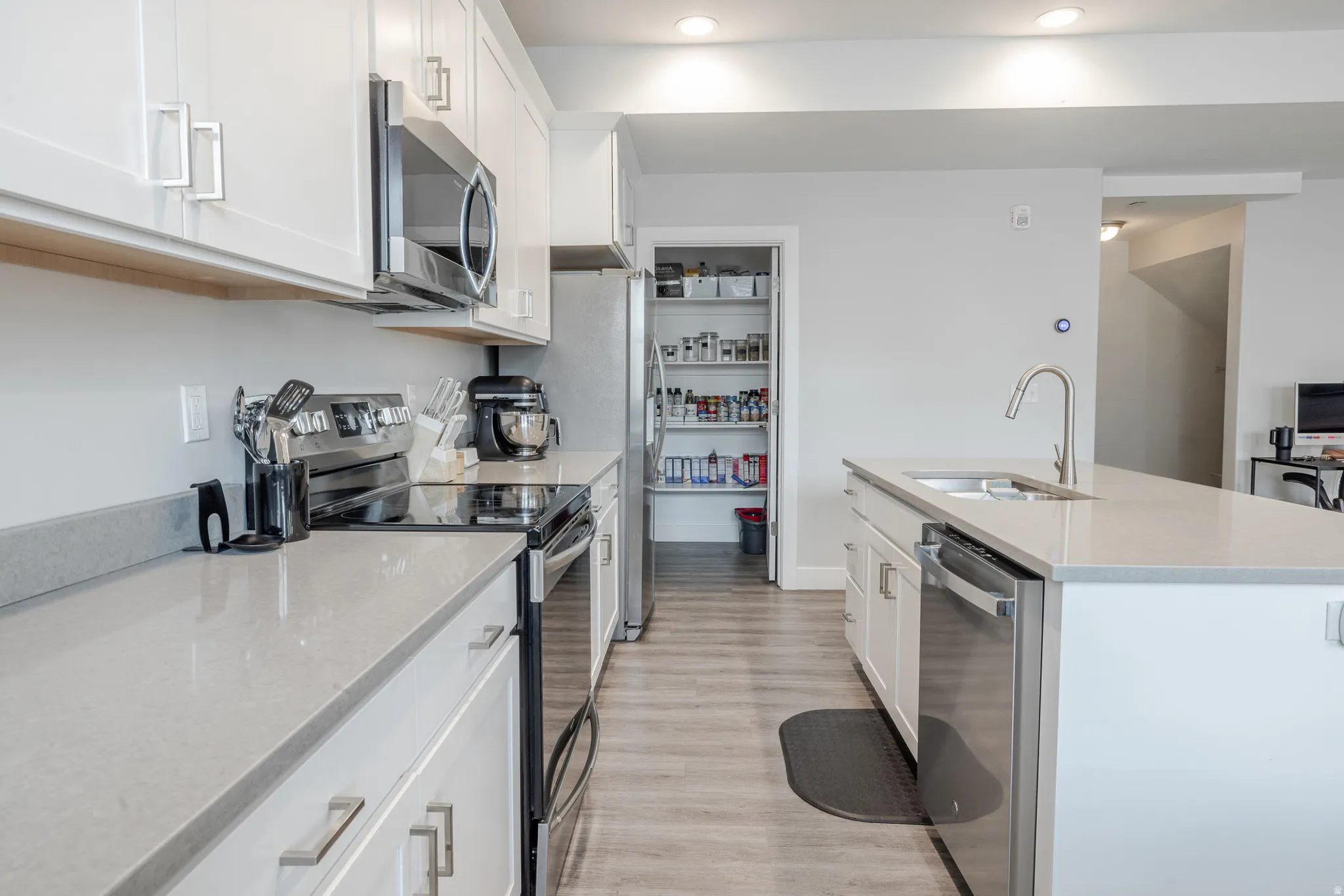 Bright kitchen featuring white cabinetry, light-toned countertops, stainless steel appliances, a center island with sink, and wood-finish flooring