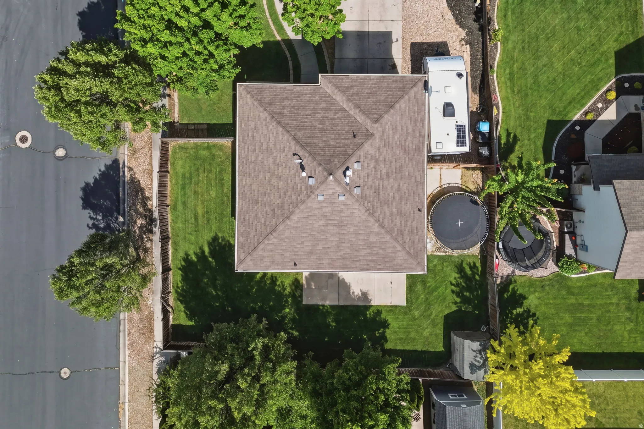 Pitched shingle roof with multiple vents and surrounding green lawns, bordered by mature trees and a paved street