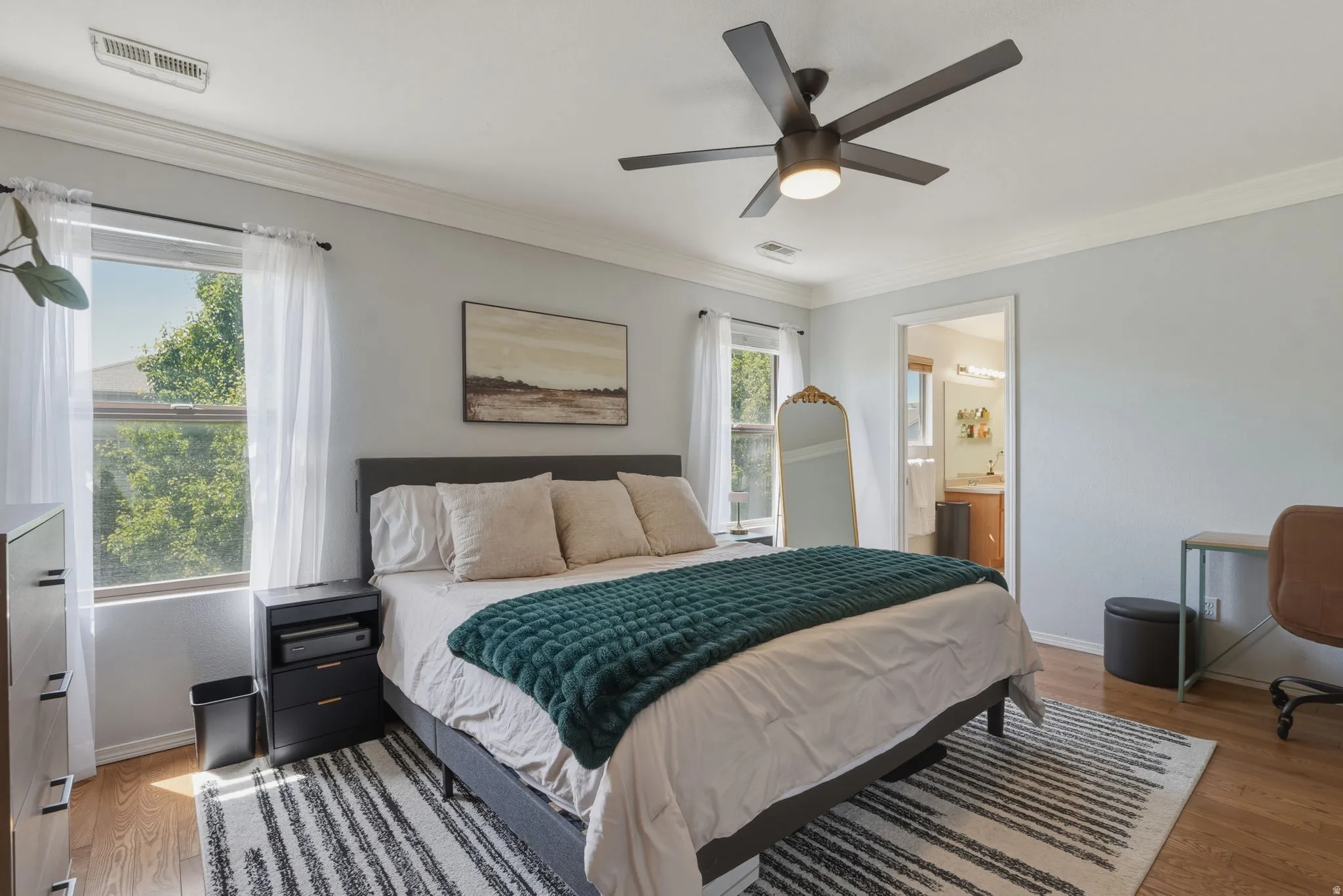 Spacious bedroom featuring wood-finish flooring, two windows with white trim, a ceiling fan with integrated lighting, and crown molding