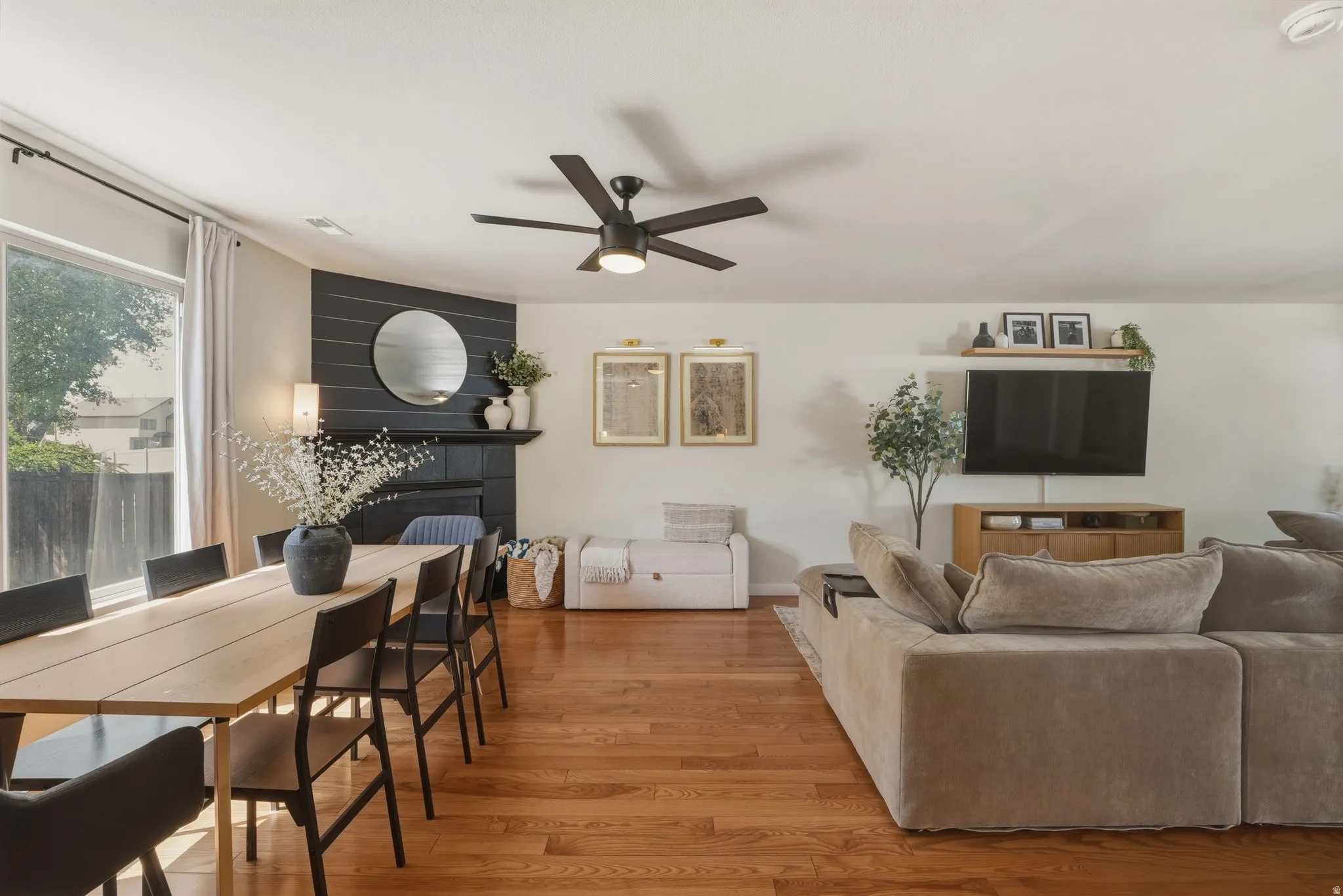 Hardwood flooring throughout a living area featuring a fireplace with black shiplap surround, a ceiling fan, and a large window