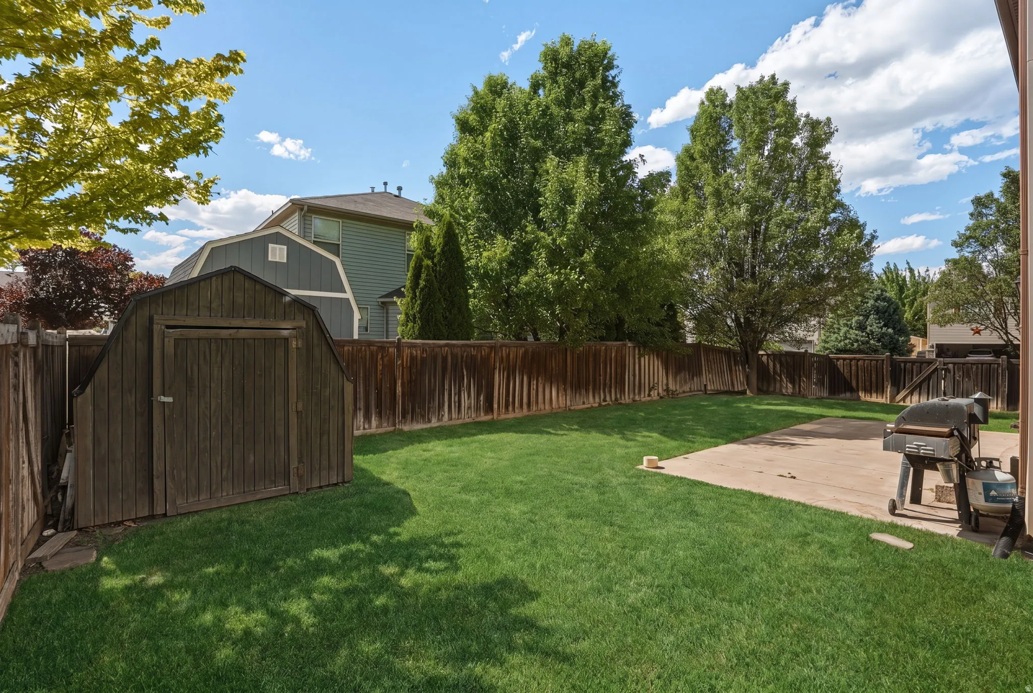 Expansive backyard featuring a lush lawn, mature trees, a wooden privacy fence, a concrete patio, and a storage shed