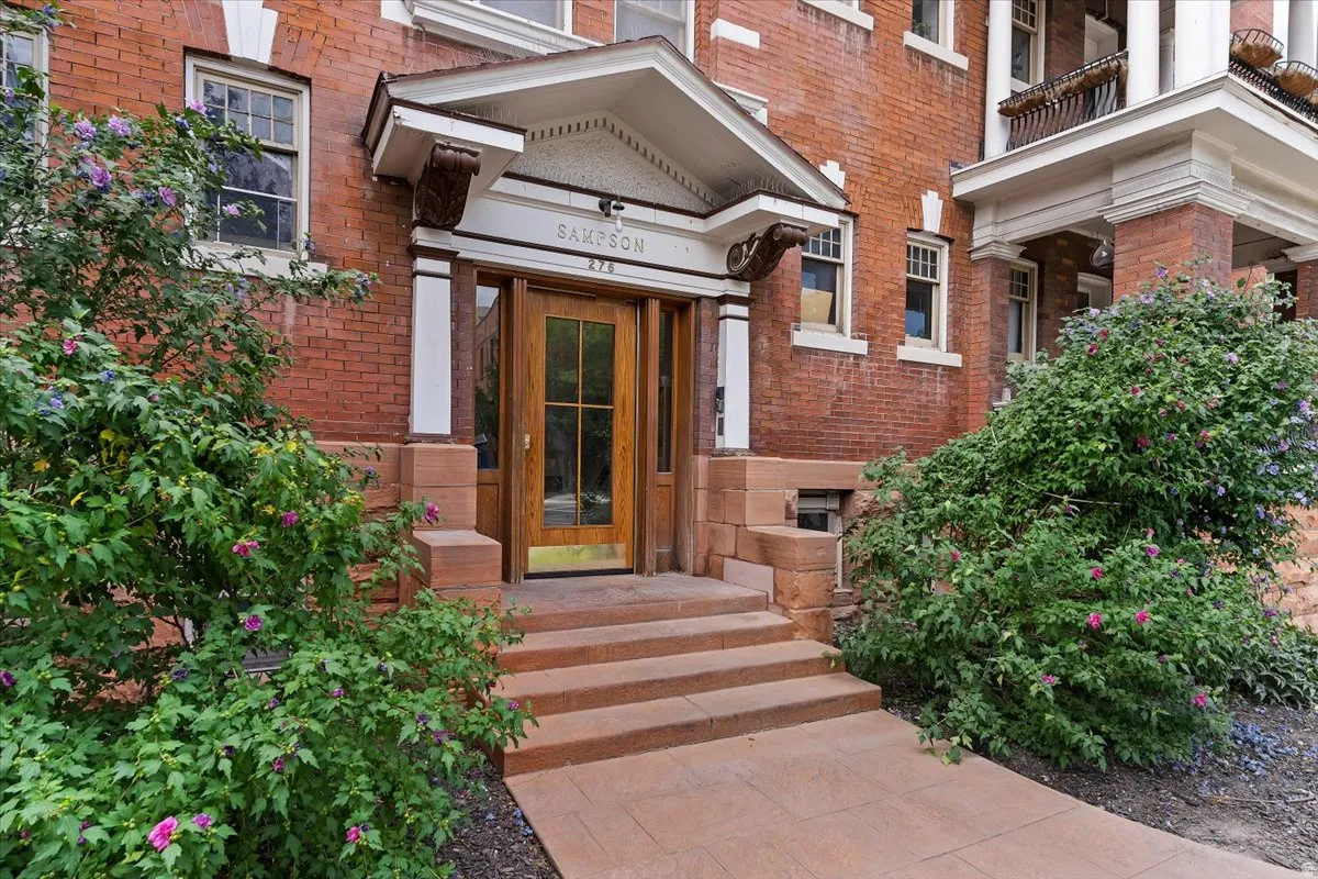 Brick facade building entrance featuring a wood-panel glass-paned door, stone steps, a covered entryway with decorative brackets, and established shrubbery