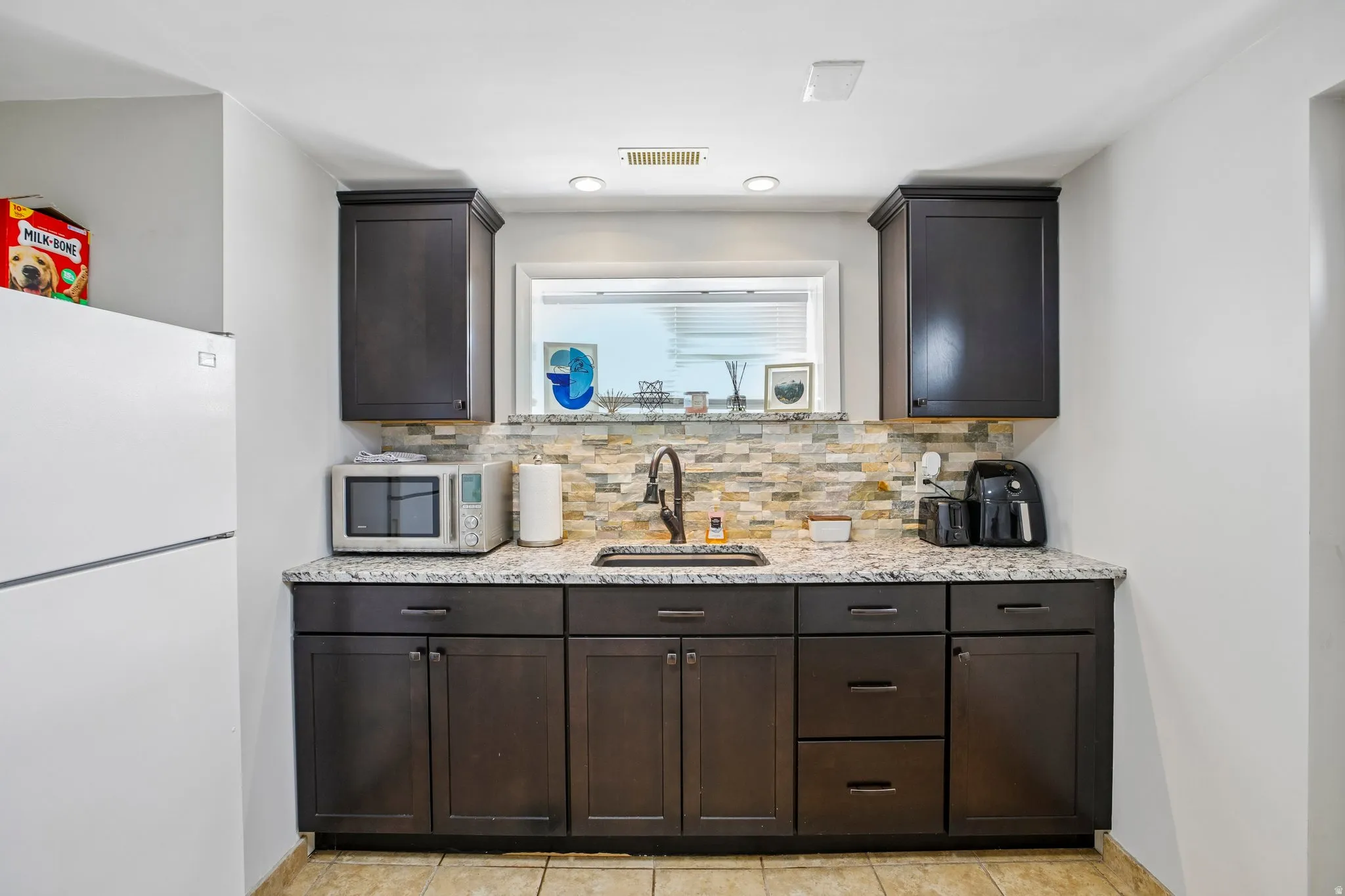 Kitchenette featuring dark wood cabinetry, a granite-finish countertop, and a stacked stone backsplash