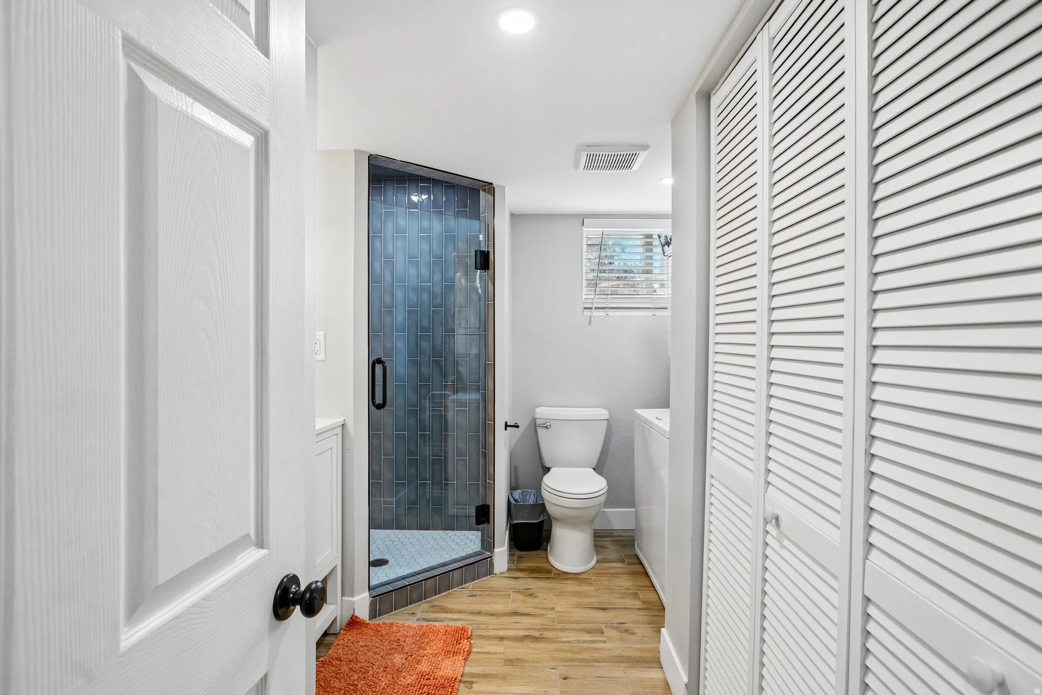 Bathroom featuring a glass-enclosed shower with dark vertical tile, wood-finish flooring, a white toilet, and louvered bi-fold closet doors