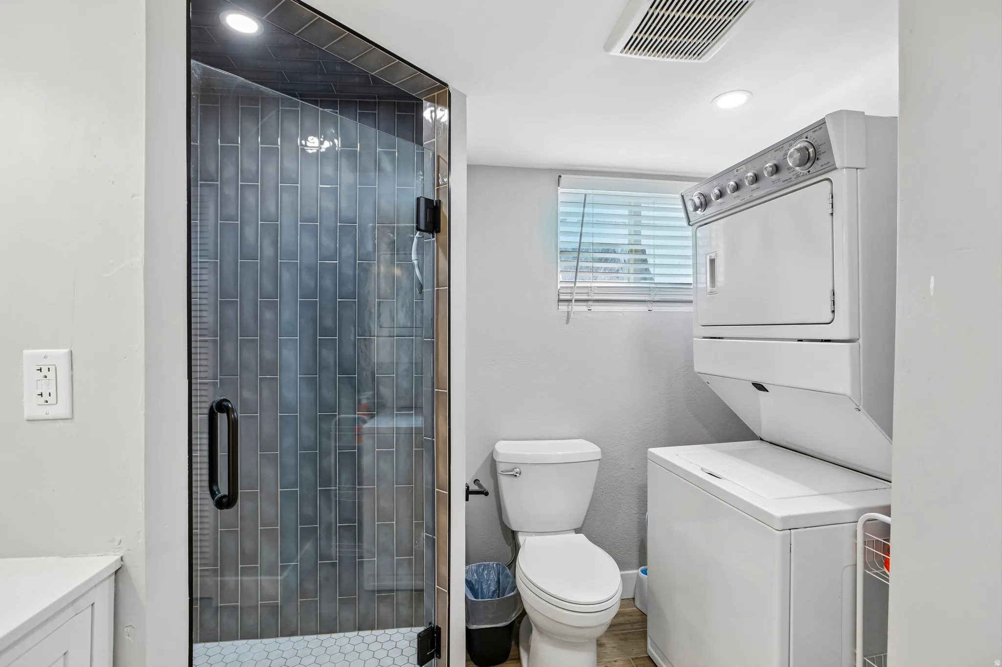 Full-size stacked washer and dryer, glass-enclosed shower with subway tile, white toilet, and recessed lighting