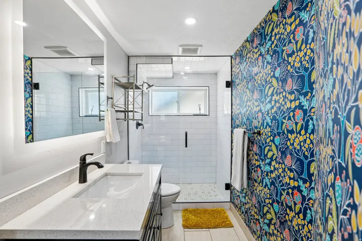 Bathroom featuring a patterned accent wall, glass-enclosed shower with subway tile, light-colored wood-finish flooring, and a vanity with a white countertop and integrated sink