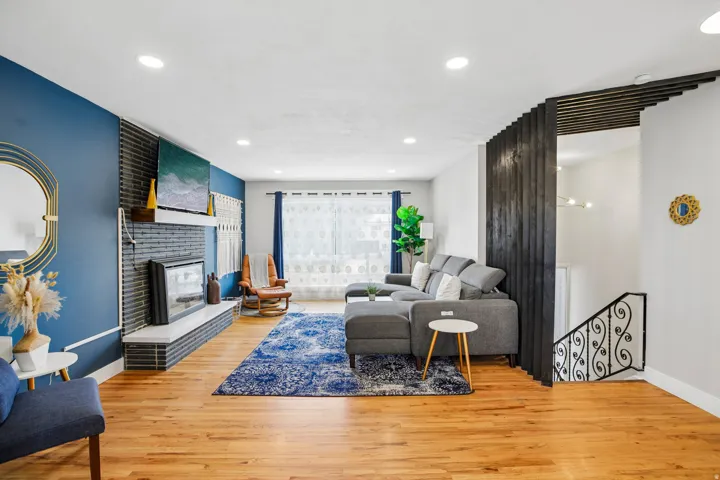 Spacious living area featuring light wood-finish flooring, recessed lighting, a modern fireplace with a black brick surround and white mantle, and a decorative wood-slat room divider