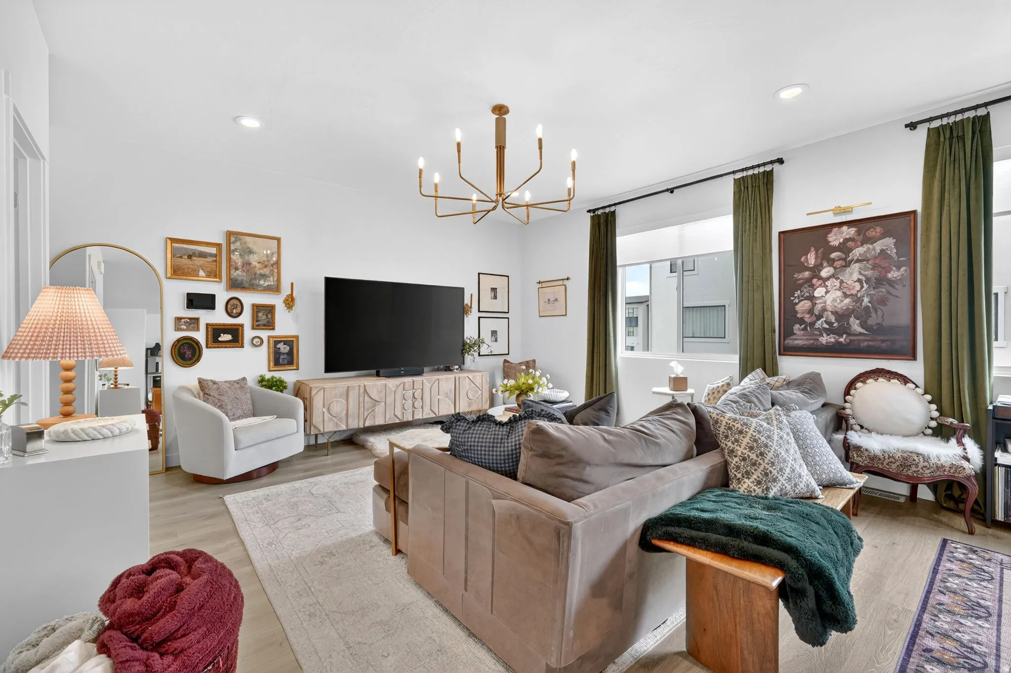 Open-concept living space featuring wood-finish flooring, a contemporary chandelier, large windows with full-length drapery, and a decorative media console