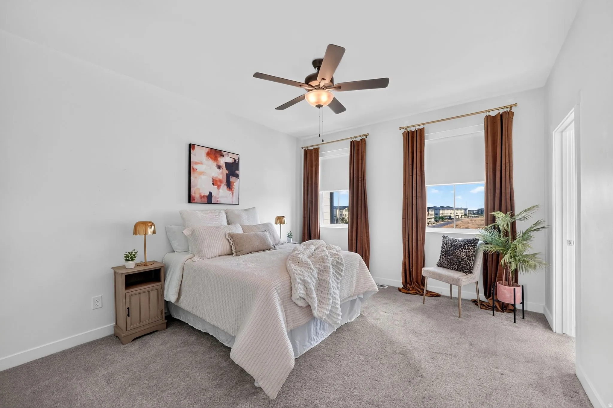 Carpeted bedroom featuring white walls, a ceiling fan with integrated lighting, two windows with roller shades and drapery rods, and a white interior door