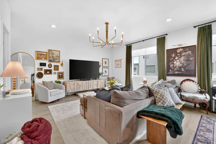 Open-concept living space featuring wood-finish flooring, a contemporary chandelier, large windows with full-length drapery, and a decorative media console