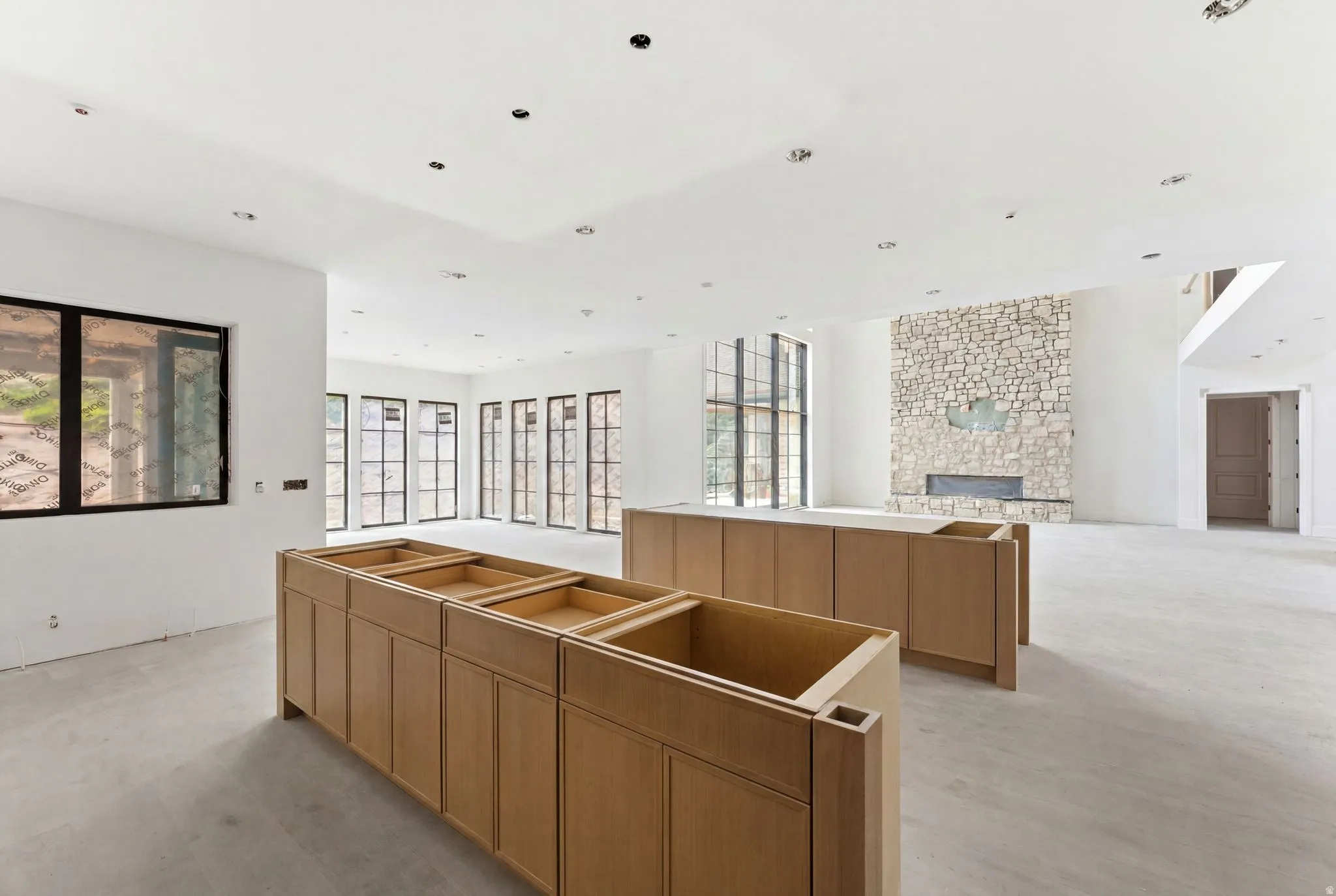 Spacious open-concept interior featuring light wood-finish cabinetry, a stone accent wall with a built-in fireplace, recessed ceiling lighting, and multiple black-framed windows providing abundant natural light