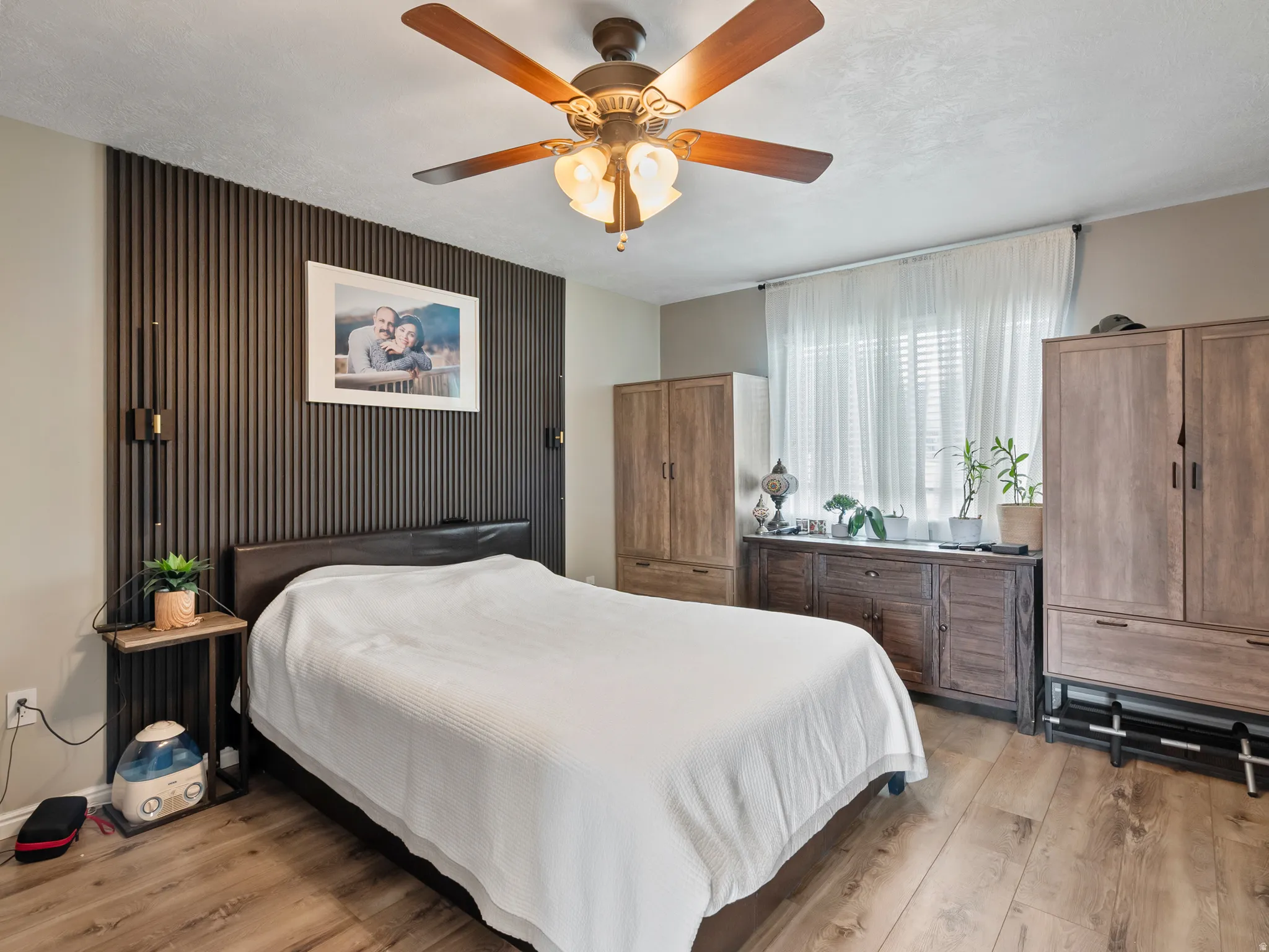 Bedroom featuring a dark slat-panel accent wall with wall-mounted sconces, wood-finish flooring, a ceiling fan with integrated lighting, and a window with blinds and sheer curtains