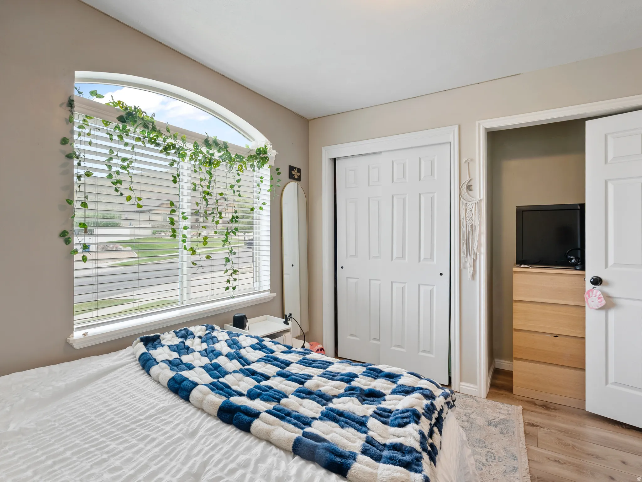 Bedroom featuring an arched window with blinds, light tan wall color, wood-finish flooring, a white bi-fold closet door, and a white interior door