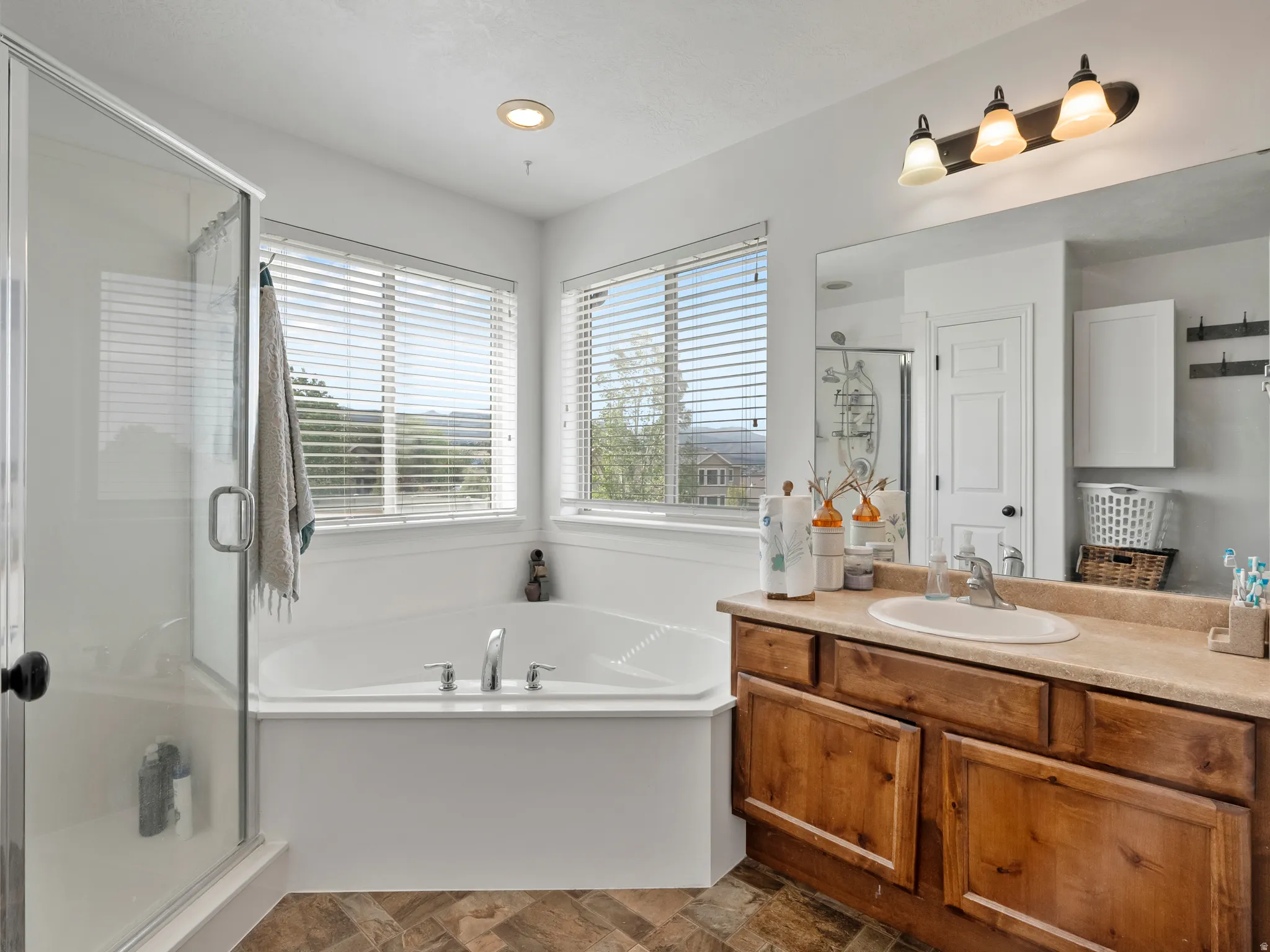 Bathroom featuring a corner soaking tub, an adjacent glass-enclosed shower, wood-finish flooring, a vanity with a light-toned countertop, and a vanity light fixture
