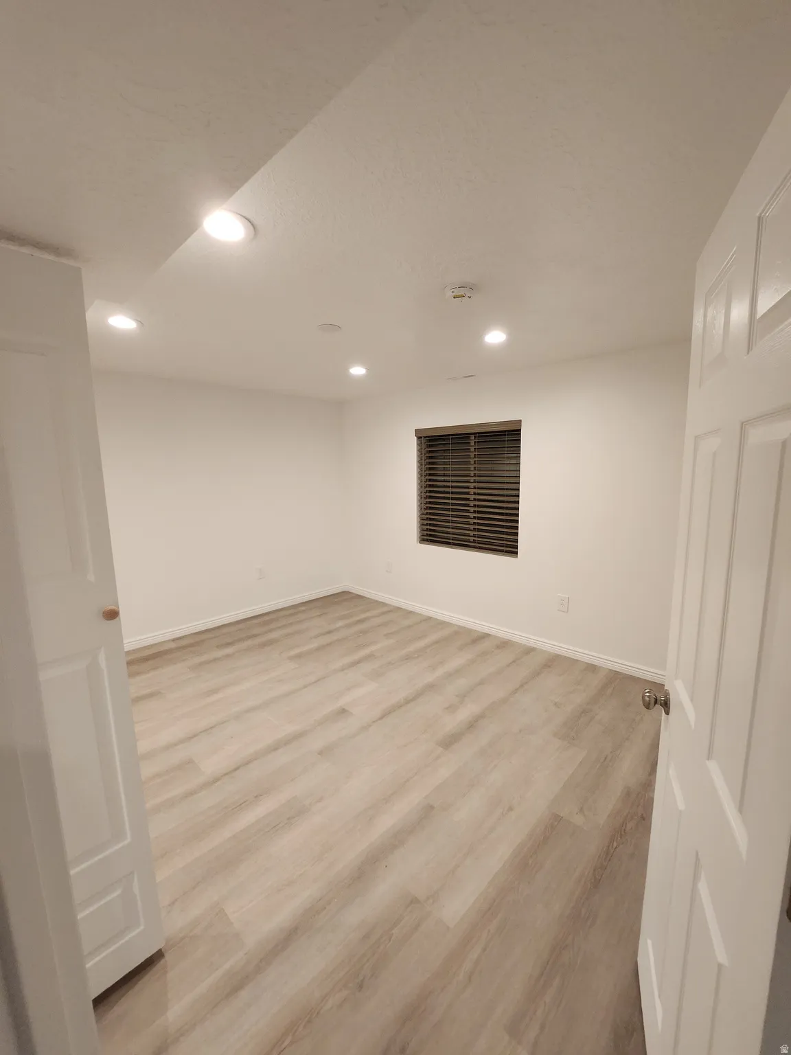 Spacious room featuring wood-finish flooring, recessed lighting, white walls, a window with horizontal blinds, and a white paneled door