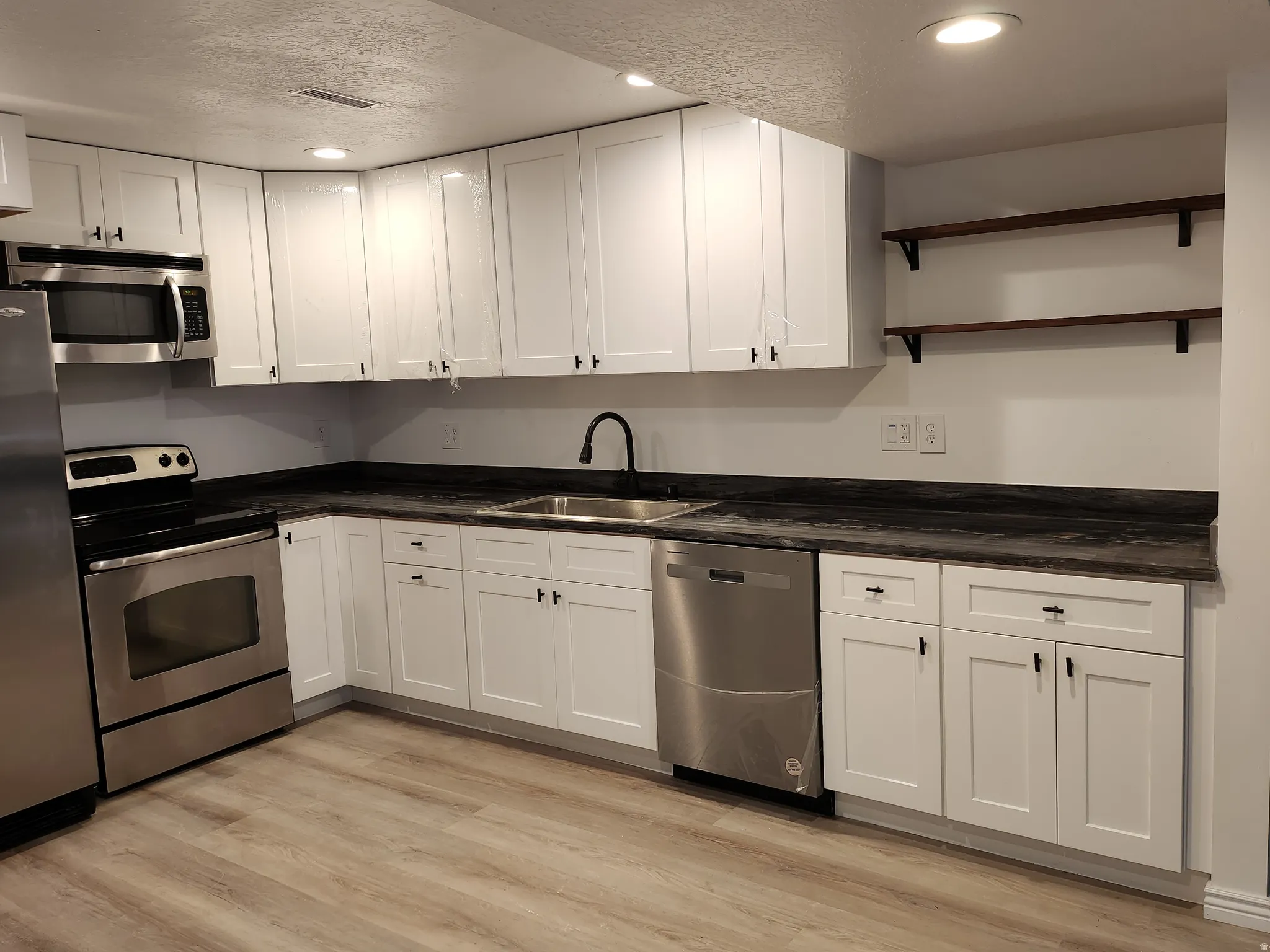 Kitchen featuring white shaker-style cabinetry, dark countertops, stainless steel appliances, and wood-finish flooring