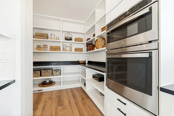 Pantry featuring extensive built-in shelving, black countertops, wood-finish flooring, and a stainless steel double oven appliance stack