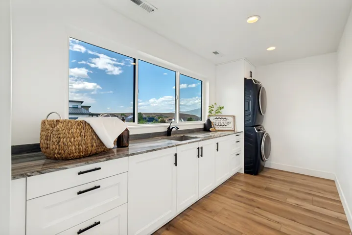Laundry room featuring a wide window, white cabinetry with black hardware, an integrated sink, stone-look countertop, and wood-finish flooring
