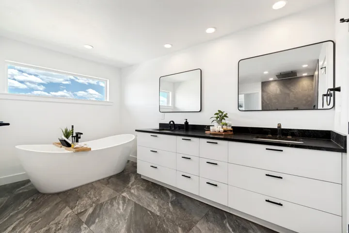 Spacious bathroom featuring a freestanding soaking tub, a dual vanity with ample storage, dark countertops, and large format floor tiling