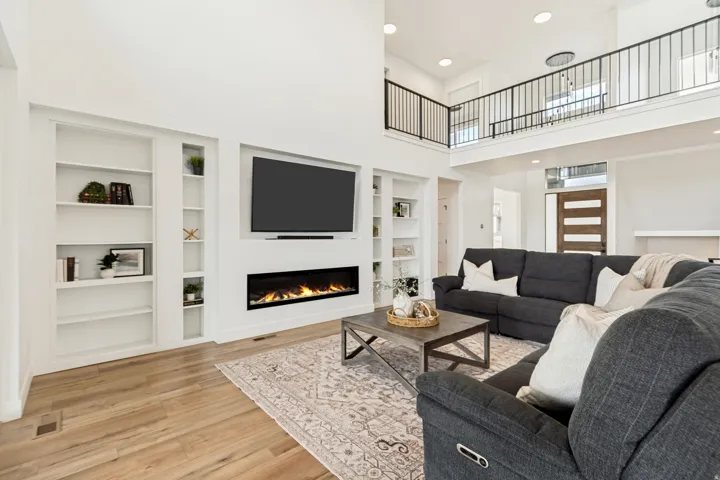 Two-story living area featuring a wall-mounted electric fireplace, built-in shelving, wood-finish flooring, and a second-level balcony with black iron railing