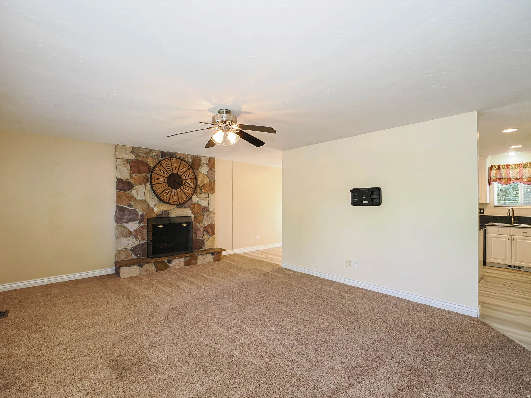 Stone-surround fireplace with a hearth, carpet flooring, a ceiling fan, and a partial view of a kitchen with light cabinetry and dark countertops