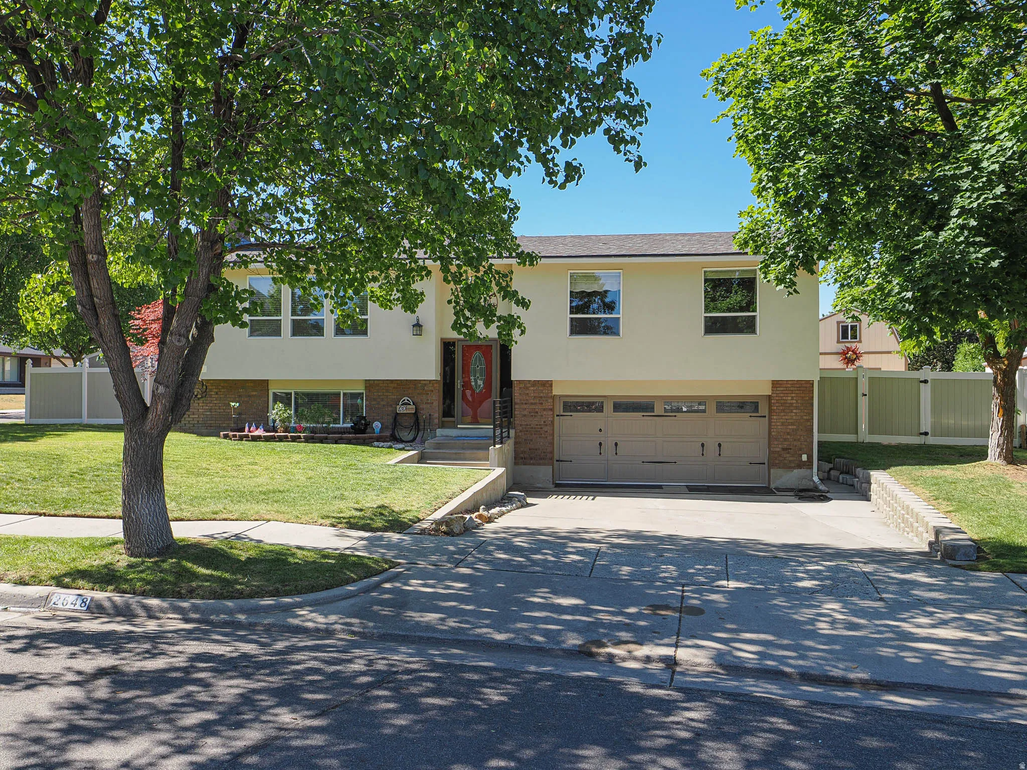 Contemporary split-level residence featuring a two-car garage, light-colored stucco and brick facade, a red entry door, and a manicured front lawn with mature trees