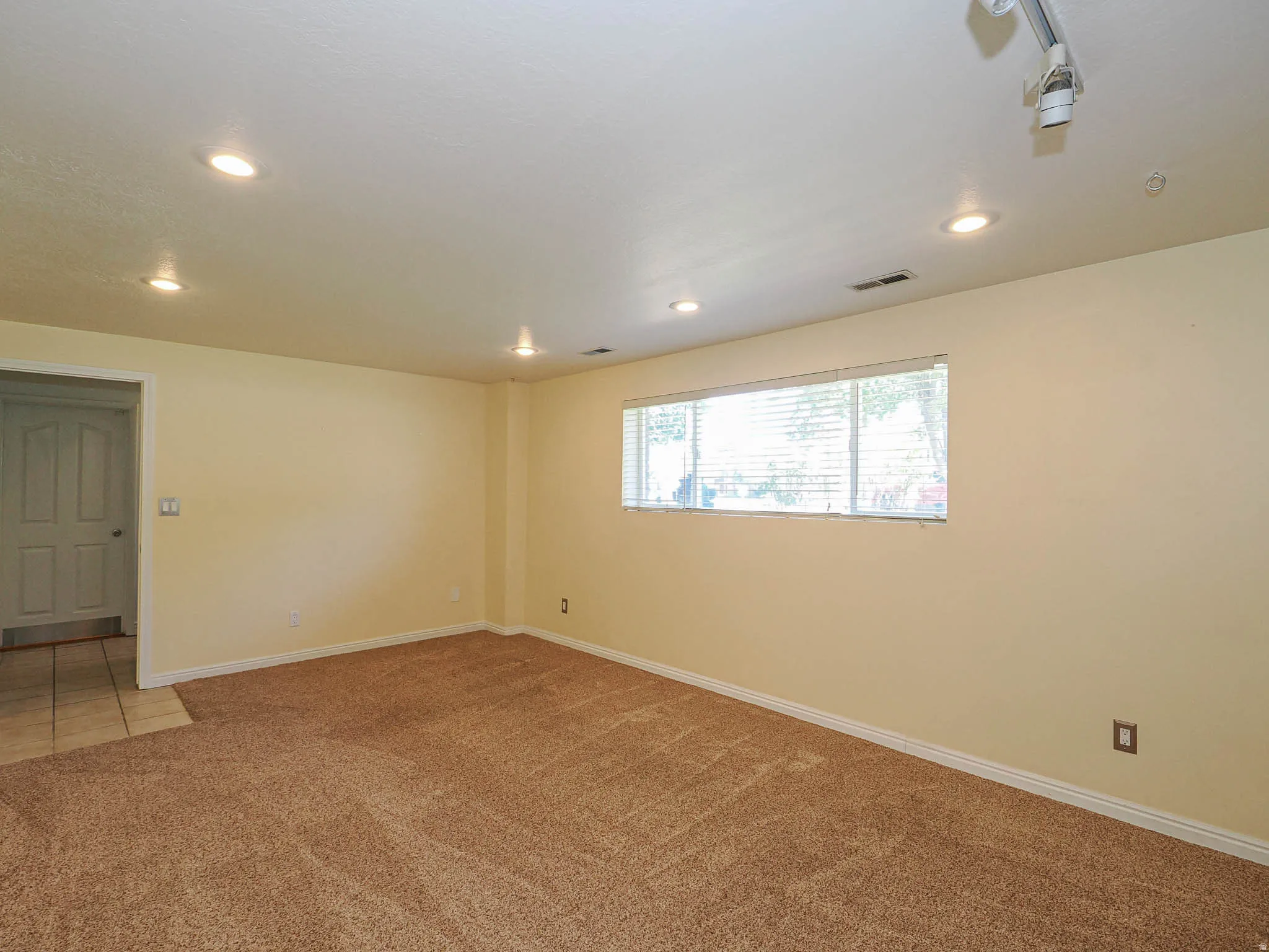 Spacious room featuring neutral carpeting, recessed ceiling lighting, and a multi-pane window with horizontal blinds