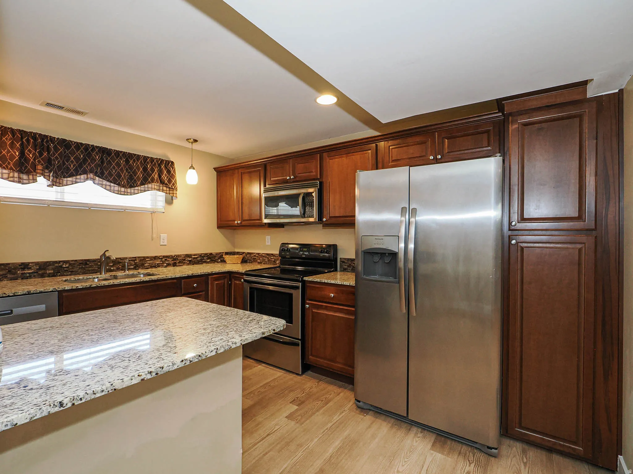 Kitchen featuring granite countertops, wood-finish flooring, stainless steel appliances, dark wood cabinetry, and a mosaic tile backsplash
