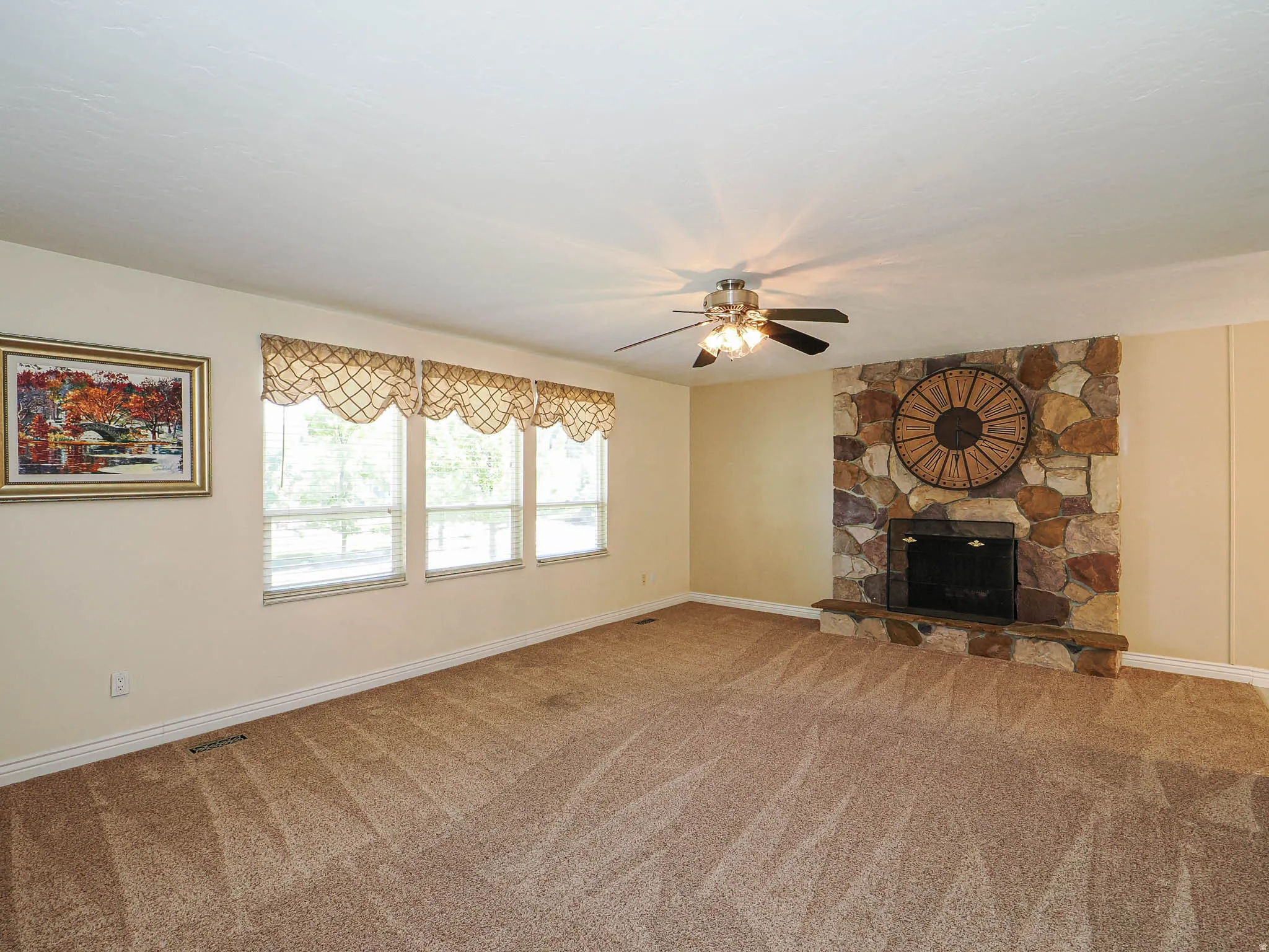 Spacious room featuring a stone-clad fireplace, carpet flooring, multiple windows with valances, and a ceiling fan with integrated lighting