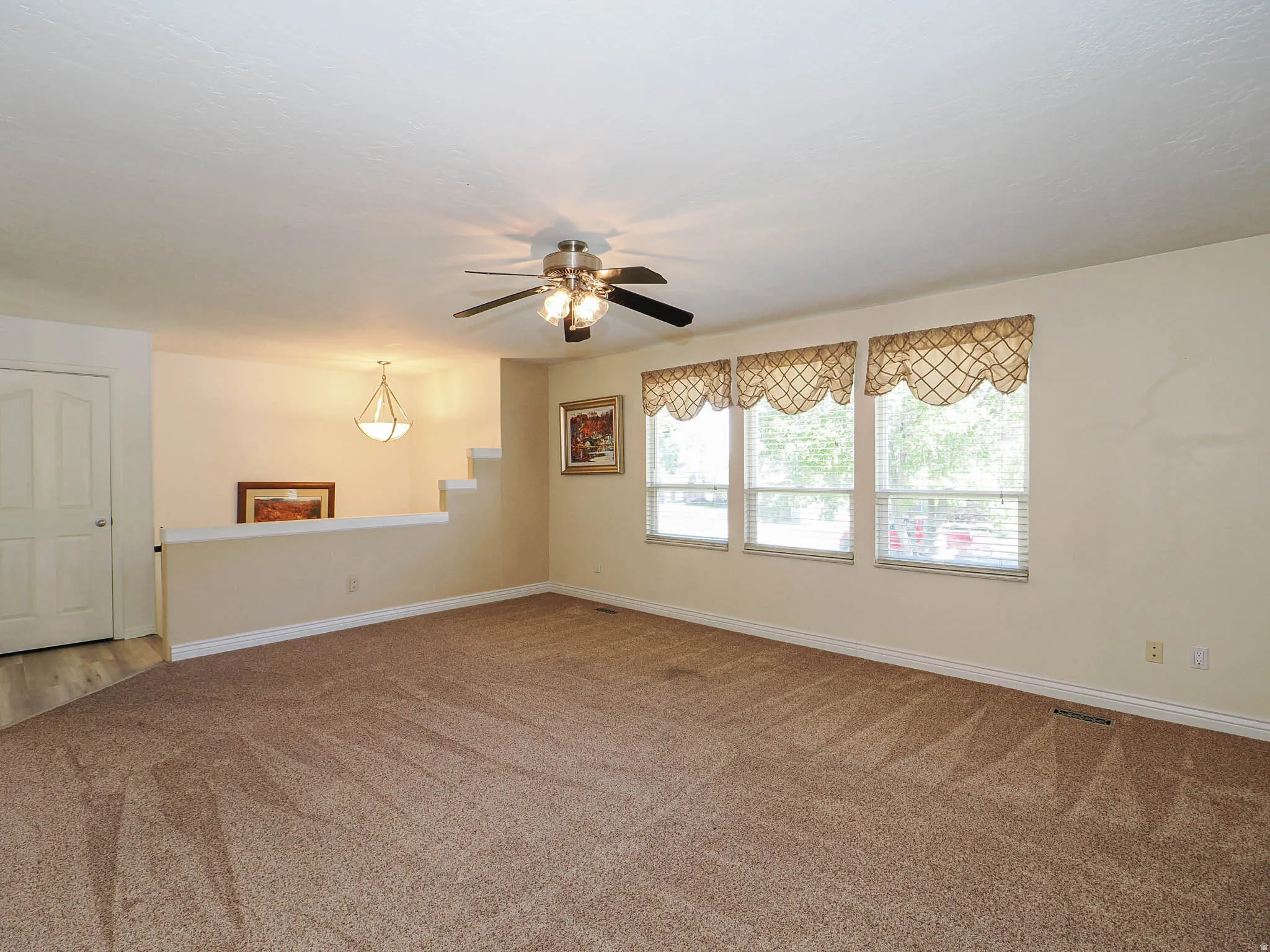 Carpeted living area featuring multiple windows with blinds, a ceiling fan with integrated lighting, and a partial wall with a wood-finish floor beyond