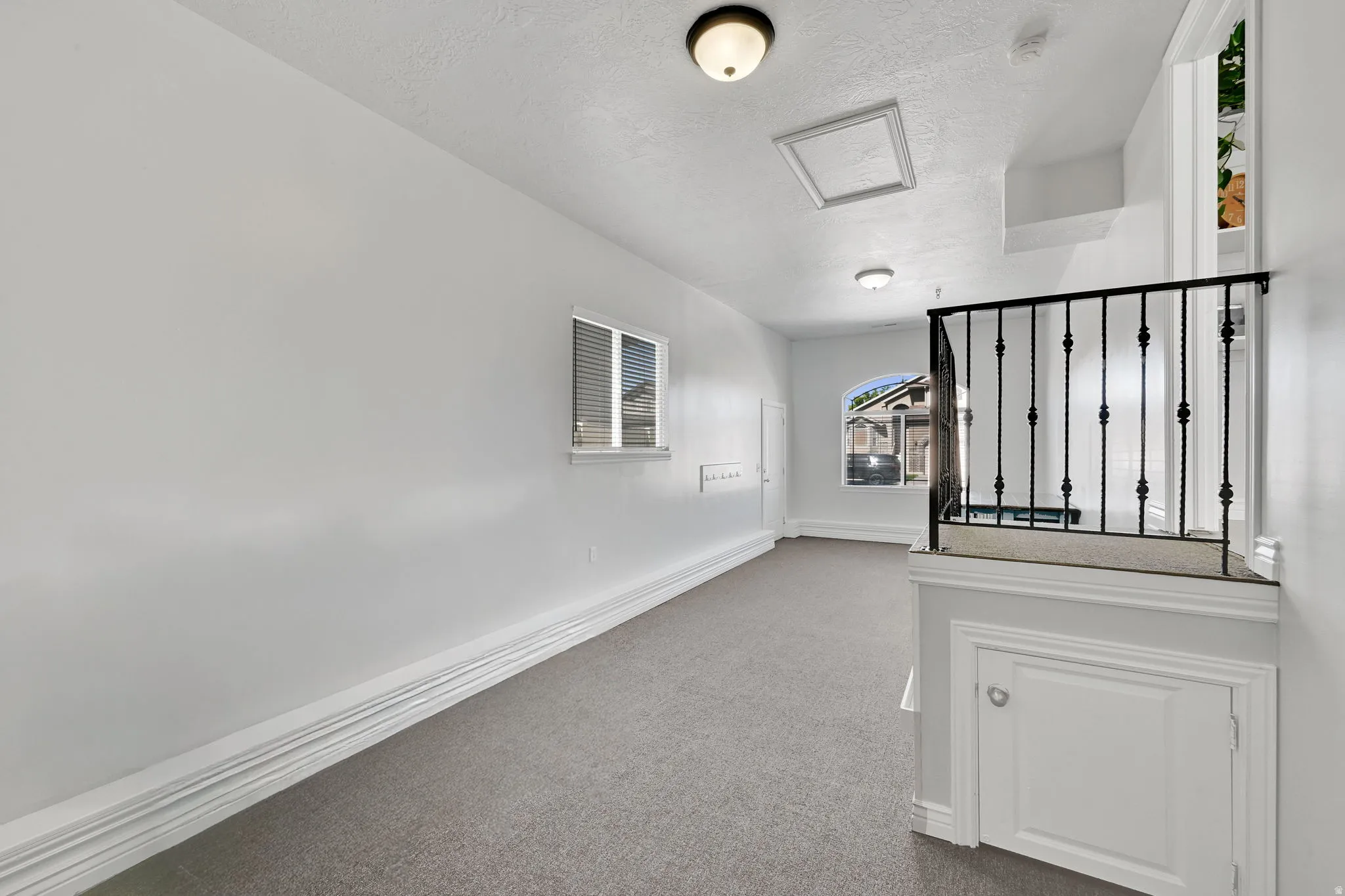 Carpeted open area featuring white walls, baseboard molding, a window with blinds, and a black wrought iron railing