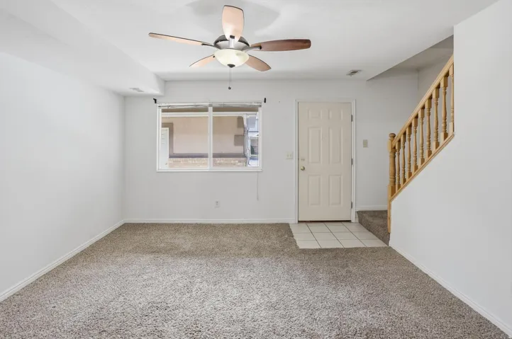 Carpeted living space featuring a ceiling fan with integrated lighting, a multi-pane window, a paneled entry door, and a staircase with wood-finish railing