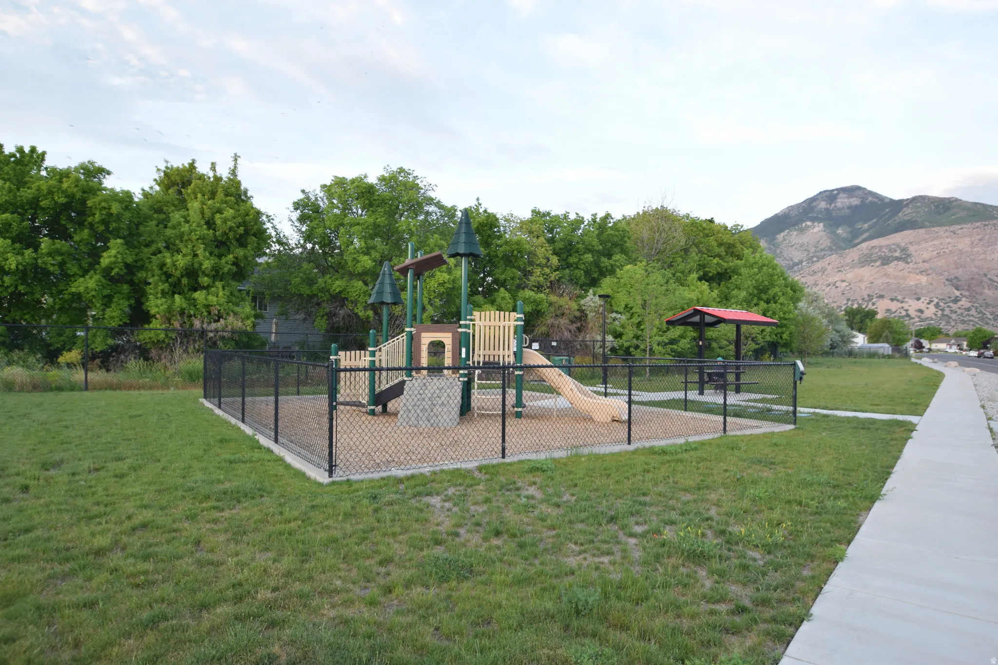 Fenced playground area featuring a play structure with slide, a separate covered picnic area, and surrounding green space