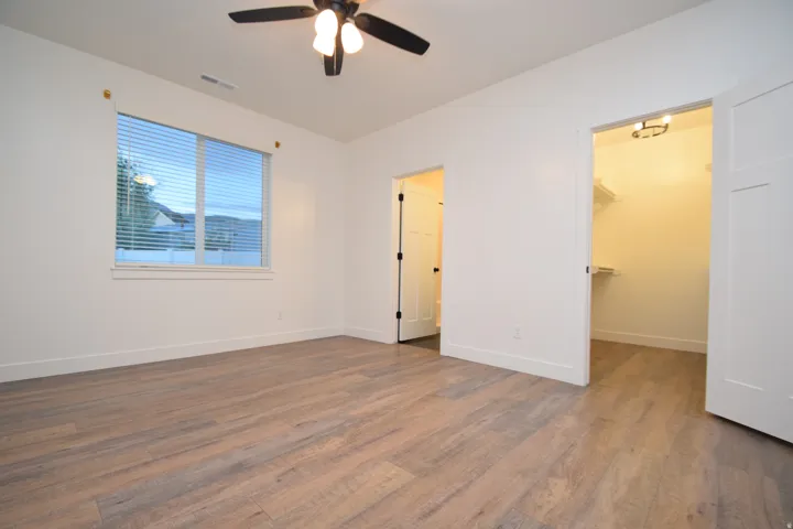 Spacious room featuring wood-finish flooring, a ceiling fan with integrated lighting, a large window with horizontal blinds, and a walk-in closet with shelving