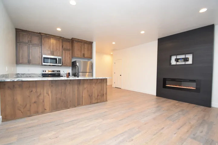Open-concept living area featuring wood-finish flooring, a contemporary linear electric fireplace with a dark shiplap accent wall, recessed lighting, a full kitchen with wood cabinetry, stainless steel appliances, and a breakfast bar with a stone-finish c
