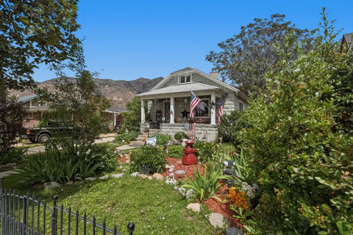 Stone facade home featuring a covered front porch with column supports, a detailed front yard with established landscaping, and a low wrought iron fence
