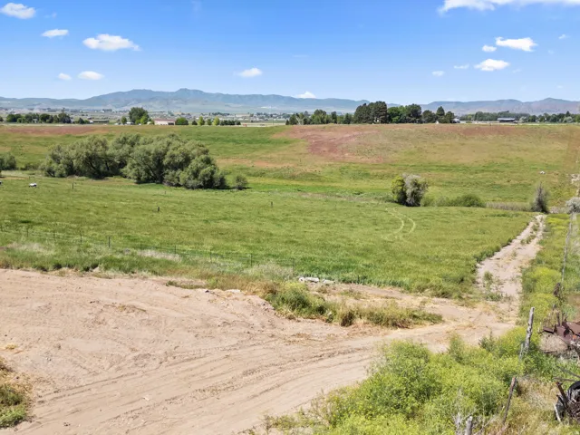 Expansive rural landscape featuring a large open field, distant mountains, scattered trees, and a dirt path