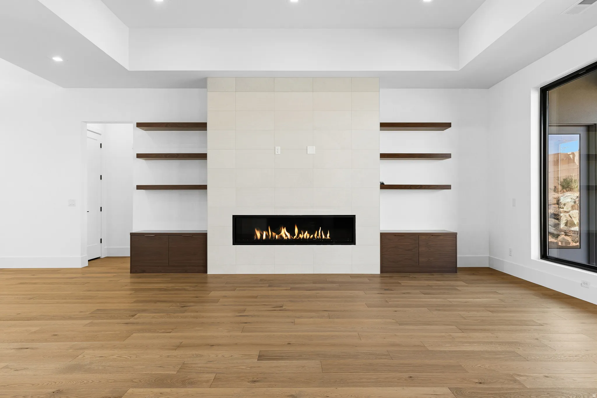 Modern living area featuring a linear fireplace with a light-toned tile surround, built-in shelving, wood-finish flooring, recessed lighting, and a large picture window